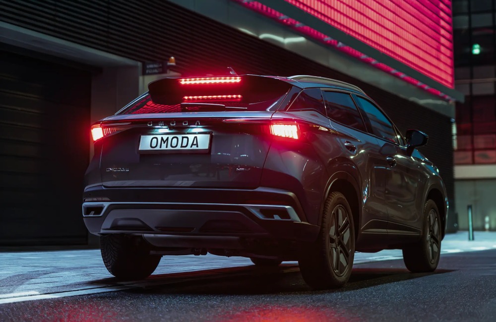 Rear shot of an OMODA 5 SHS-H, showing off the lit rear lighting and OMODA branding. The backdrop consists of modern buildings at night as the car is parked in the street.