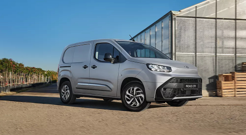 Proace City compact panel van, in silver, driving beside a greenhouse building.