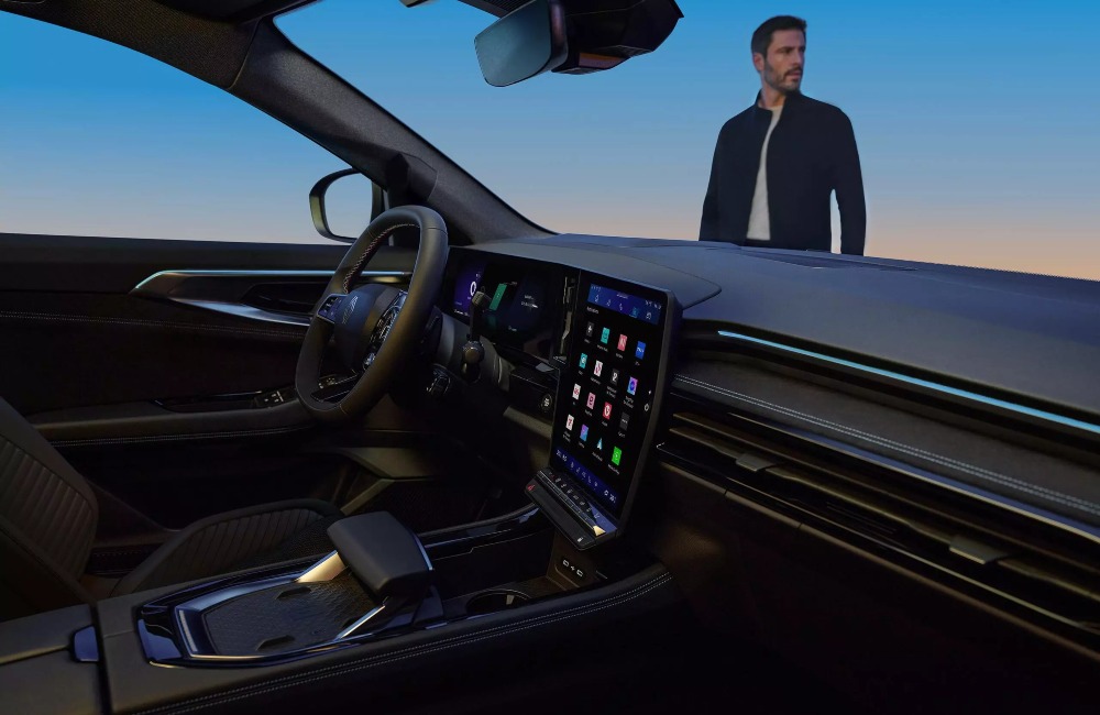 Interior shot of a new Renault Austral, with focus on the drivers cockpit and infotainment system as a whole. There is a person stood outside the car looking into the distance.