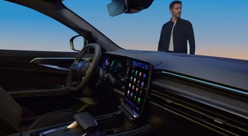 Interior shot of a new Renault Austral, with focus on the drivers cockpit and infotainment system as a whole. There is a person stood outside the car looking into the distance.