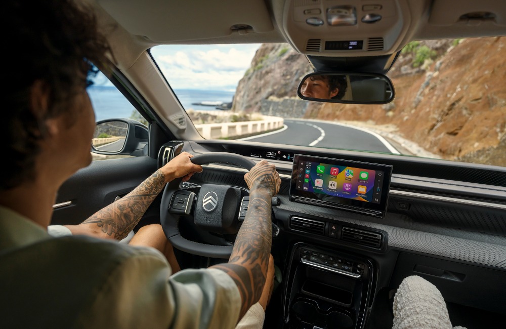Interior shot of a person driving a new Citroen C3 Aircross down a coastal road at speed. The shot focusses on the person driving and shows off the infotainment system