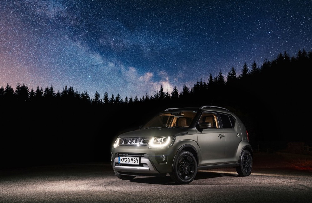 Angled shot of a Suzuki Ignis at night with a star-filled sky background. The car has its front headlights lit and is surrounded by dark forest, making it stand out with the night sky.
