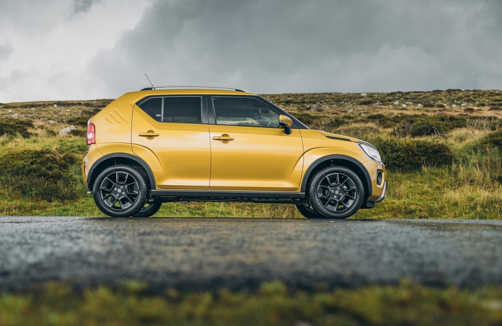 Side profile of a yellow Suzuki Ignis parked on a wet countryside road.