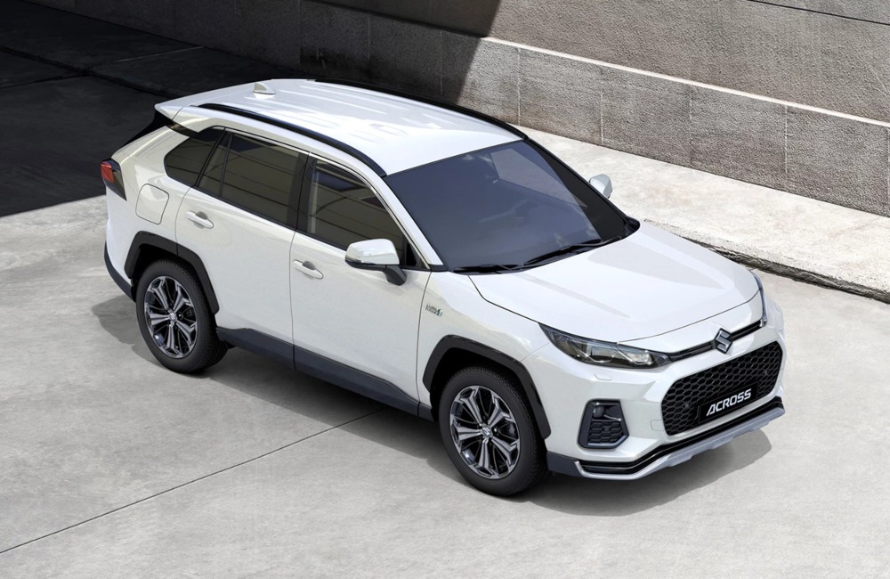 Angled top-down view of a new Suzuki Across, showing off the roof and front styling. The car is parked on a concrete road outside a concrete building.