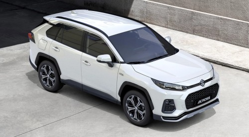 Angled top-down view of a new Suzuki Across, showing off the roof and front styling. The car is parked on a concrete road outside a concrete building.
