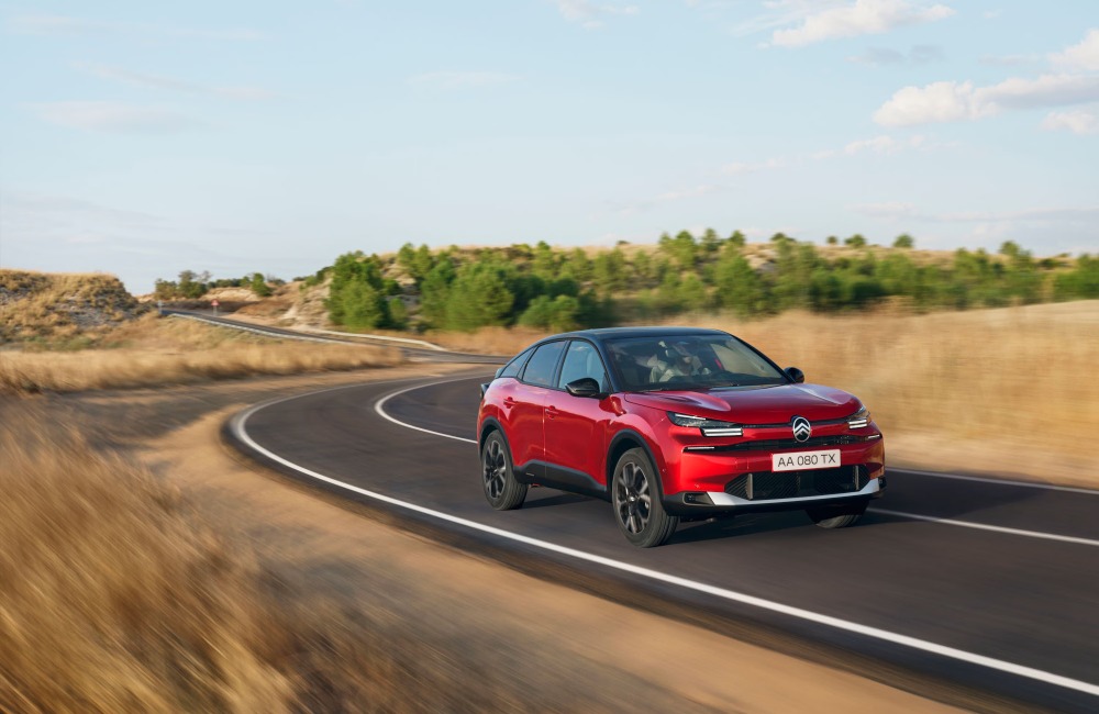 Citroen C4 driving at speed down a windy country-side road, surrounded by brown grass and green trees.