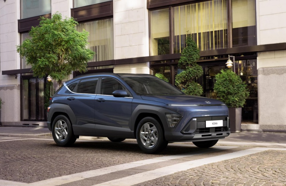 Angled shot of a Hyundai Kona, showing off the front and side design. The car is parked on a cobbled road outside a modern building.