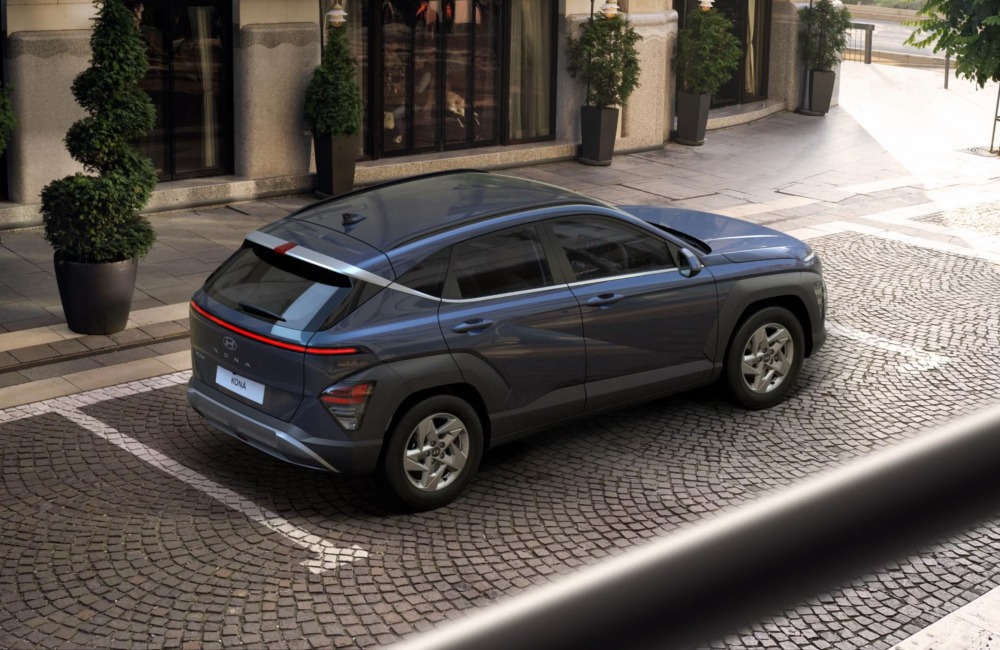 Angled top down view of a Hyundai KONA, showing off the tailgate, side, and roof. The car is parked on a cobbled road outside a modern building.