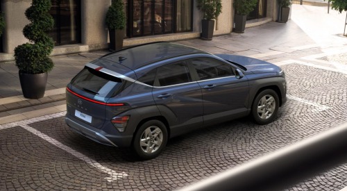 Angled top down view of a Hyundai KONA, showing off the tailgate, side, and roof. The car is parked on a cobbled road outside a modern building.
