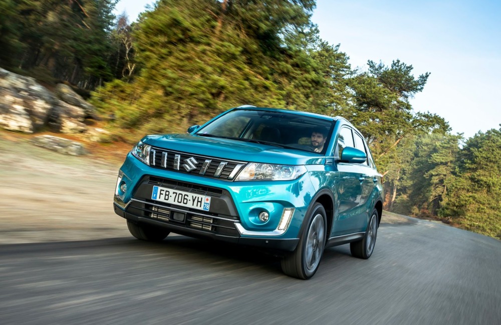 Angled front shot of a used Suzuki Vitara driving at speed down a country road surrounded by trees.