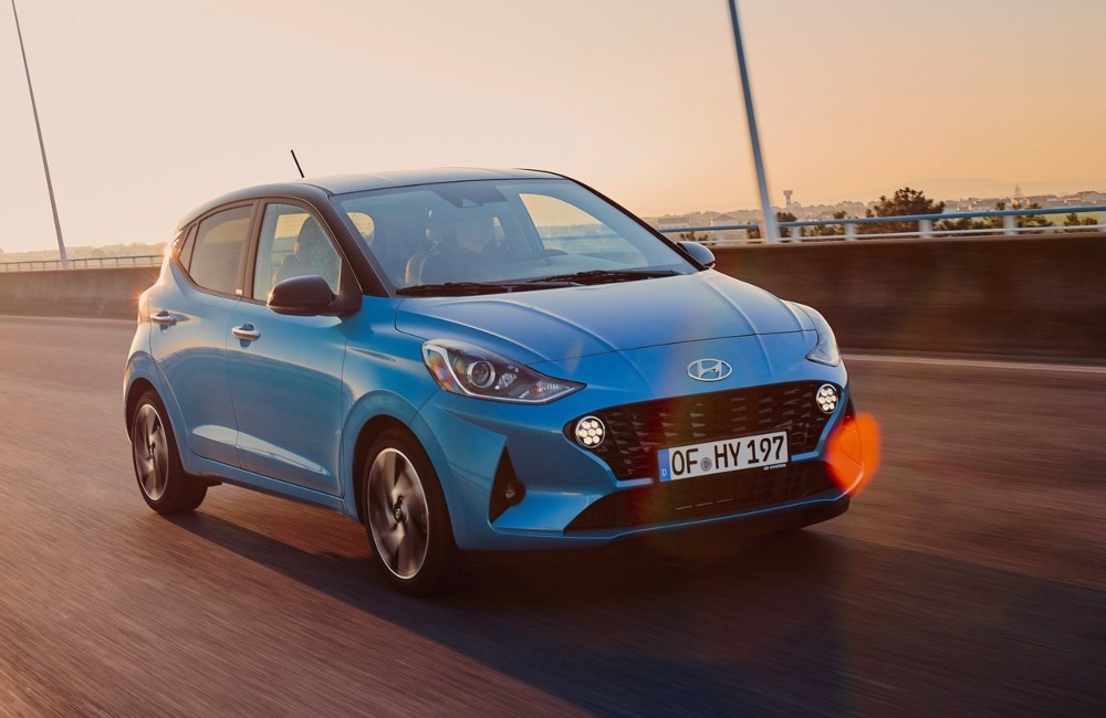 Blue Hyundai i10 driving at speed over a large bridge with a sunset backdrop.