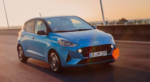 Blue Hyundai i10 driving at speed over a large bridge with a sunset backdrop.