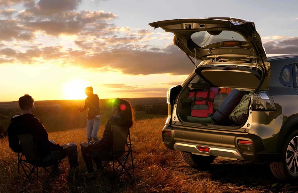 Three people enjoy the sunset next to a used Suzuki car with the boot open and camping gear inside.