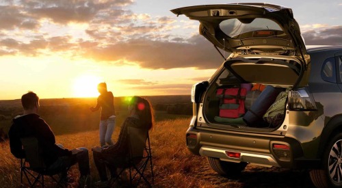 Three people enjoy the sunset next to a used Suzuki car with the boot open and camping gear inside.
