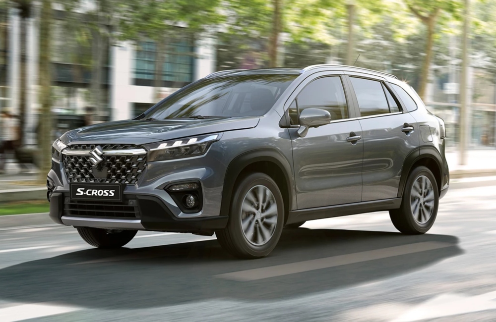 Angled front shot of a new Suzuki S-Cross travelling through a city street at speed. The car is grey and surrounded by thin trees and city buildings.