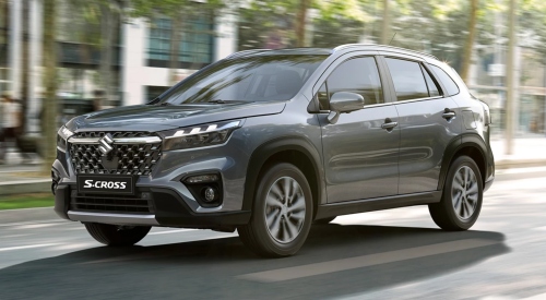 Angled front shot of a new Suzuki S-Cross travelling through a city street at speed. The car is grey and surrounded by thin trees and city buildings.