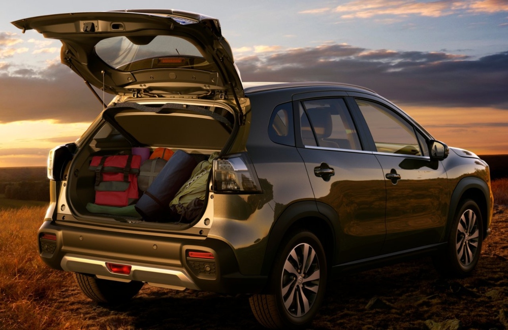 Angled rear shot of a new Suzuki S-Cross with the boot open, filled with luggage and camping equipment. The car is parked on a grassy plateau, with a sunset background.