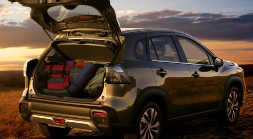 Angled rear shot of a new Suzuki S-Cross with the boot open, filled with luggage and camping equipment. The car is parked on a grassy plateau, with a sunset background.