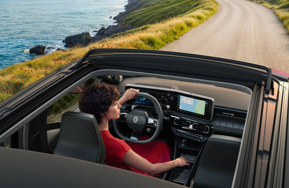 A person in a red dress driving a new Renault 4 Plein Sud down a cliffside road at speed with the canvas roof open.