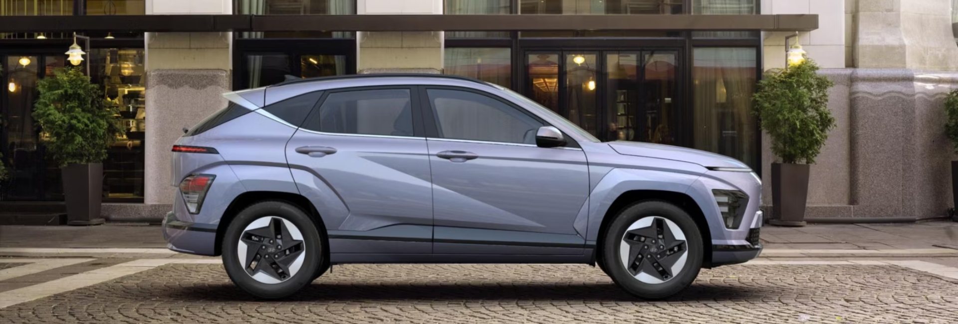 Side profile of a new Hyundai KONA Electric parked outside of a modern building on a cobbled road.