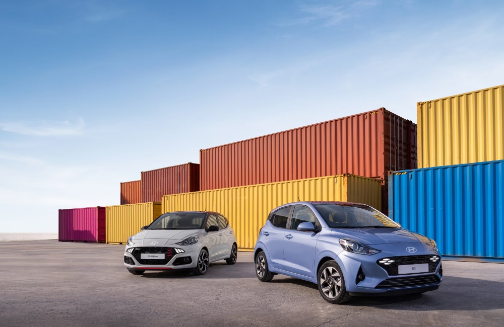 A new Hyundai i10 and i10 N Line parked next to each other in front of some shipping containers.