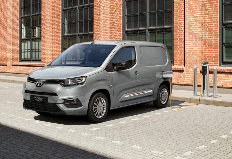 Used Toyota Proace City Electric van charging at EV station outside industrial brick building.