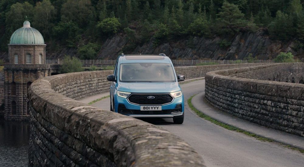 New Ford Tourneo Connect in Boundless Blue driving across a stone bridge in woodland.