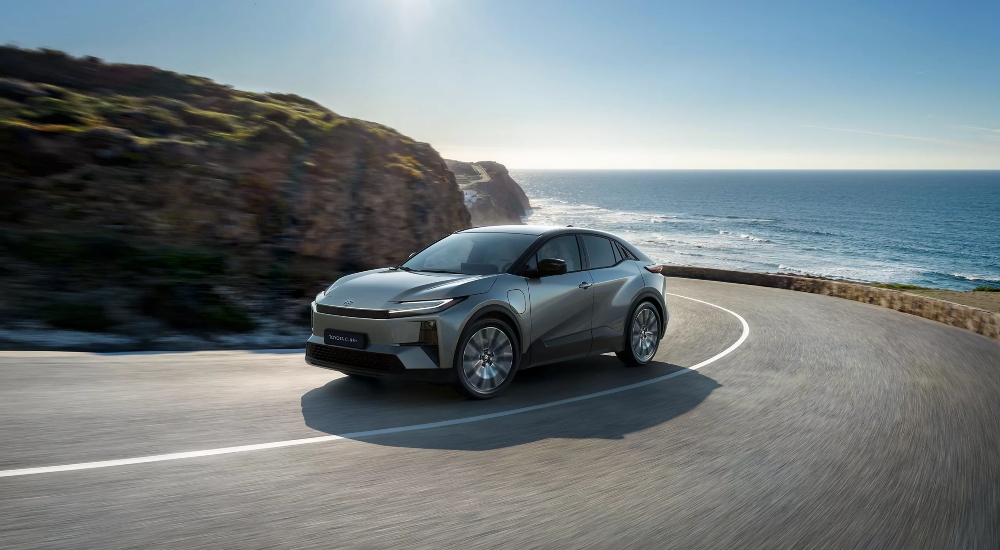 New Toyota C-HR+ driving along a coastal road, showing its coupe-SUV profile and aerodynamic design.