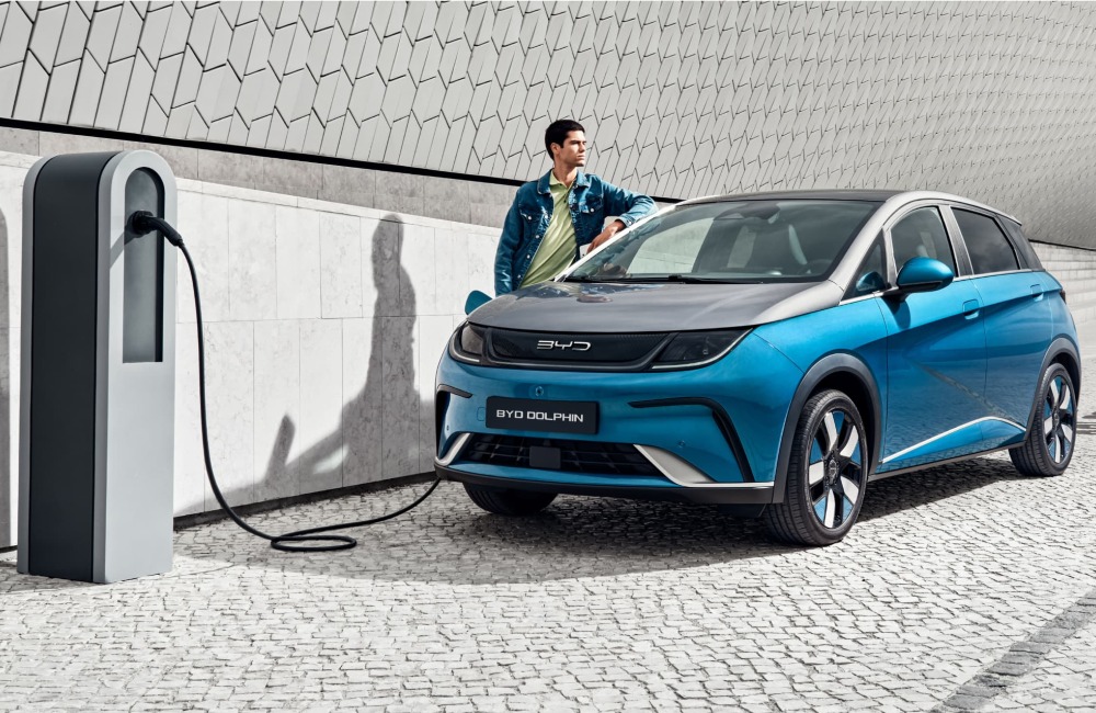 Person stood next to their parked BYD DOLPHIN, waiting for it to charge at a public charging point.