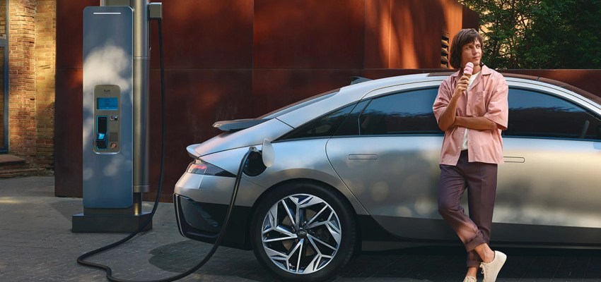 A woman leans against a silver Hyundai IONIQ 6 whilst it charges; Cost to charge an EV hub image
