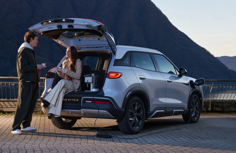 Two people conversating and drinking coffee, utilising the rear boot of a used BYD car as a seat and bench for their coffee maker whilst the tailgate is open.
