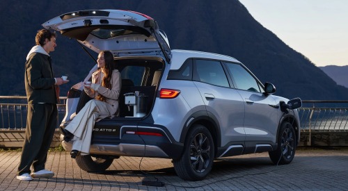 Two people conversating and drinking coffee, utilising the rear boot of a used BYD car as a seat and bench for their coffee maker whilst the tailgate is open.