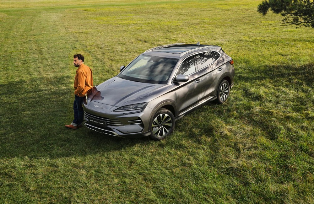 A top-angled shot of a person leaning on a used BYD car in a lush grassland.