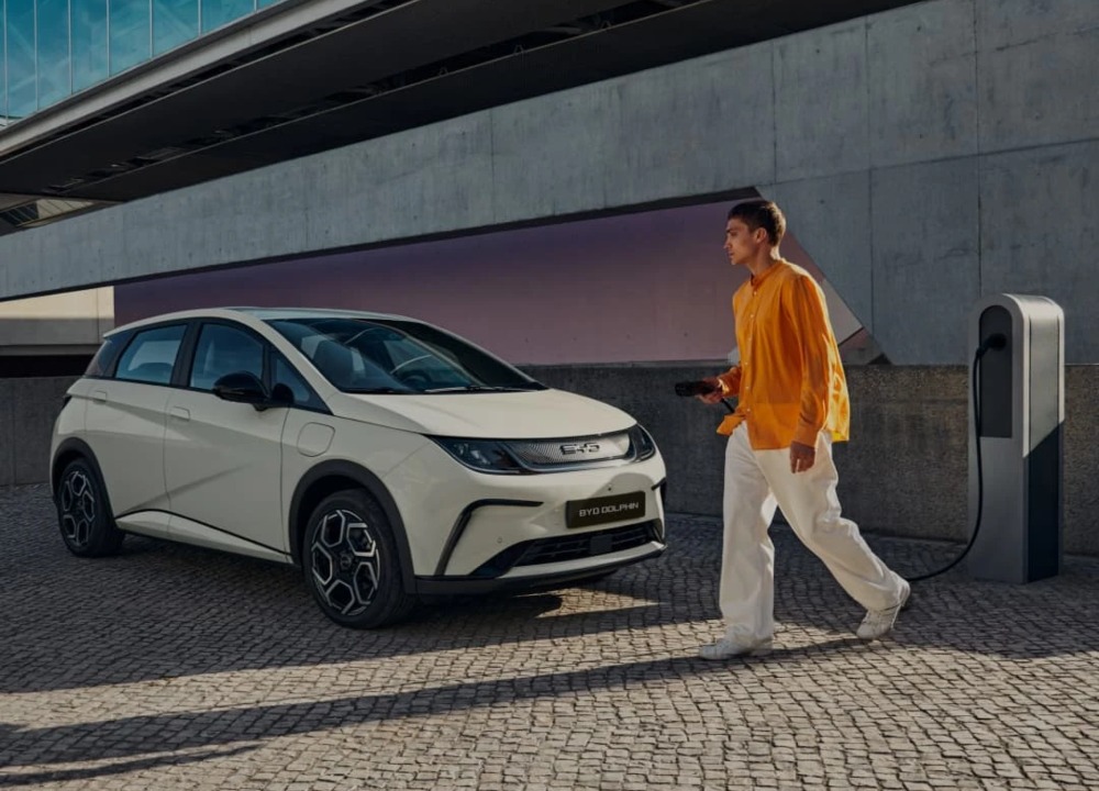 A person holding an electric car charger, walking towards a parked BYD DOLPHIN: Lease BYD