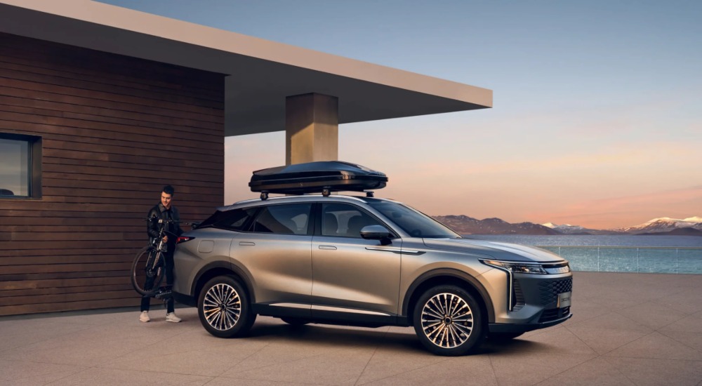 Side shot of an OMODA 9 with a roof box attached and a scenic background. A person is putting a bike on the rear of the car, showing off its capabilities.