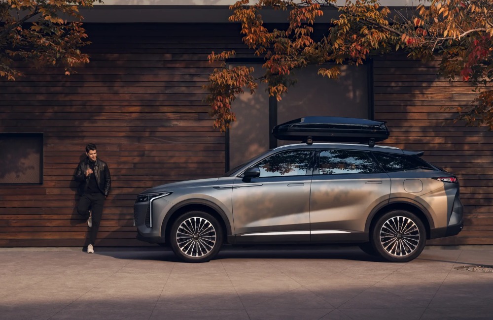 Side profile of an OMODA 9, with a person in a leather jacket leaning on a wall besides it. The car features unique alloy wheels, LED lighting, and a roofbox.