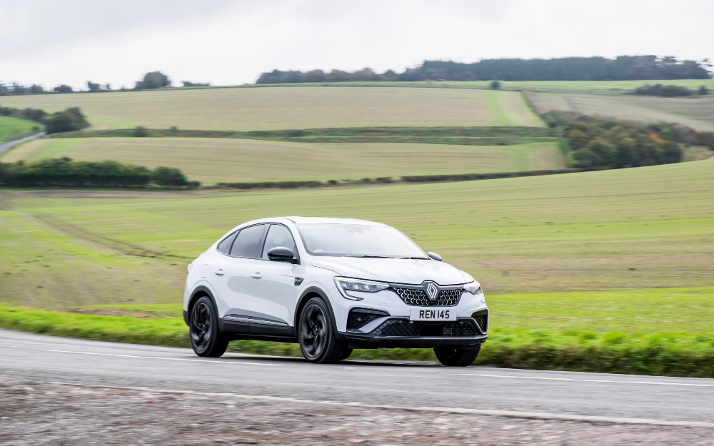 Used Renault Arkana E-Tech Renault Arkana E-Tech in white, driving on a country road with fields in the background.