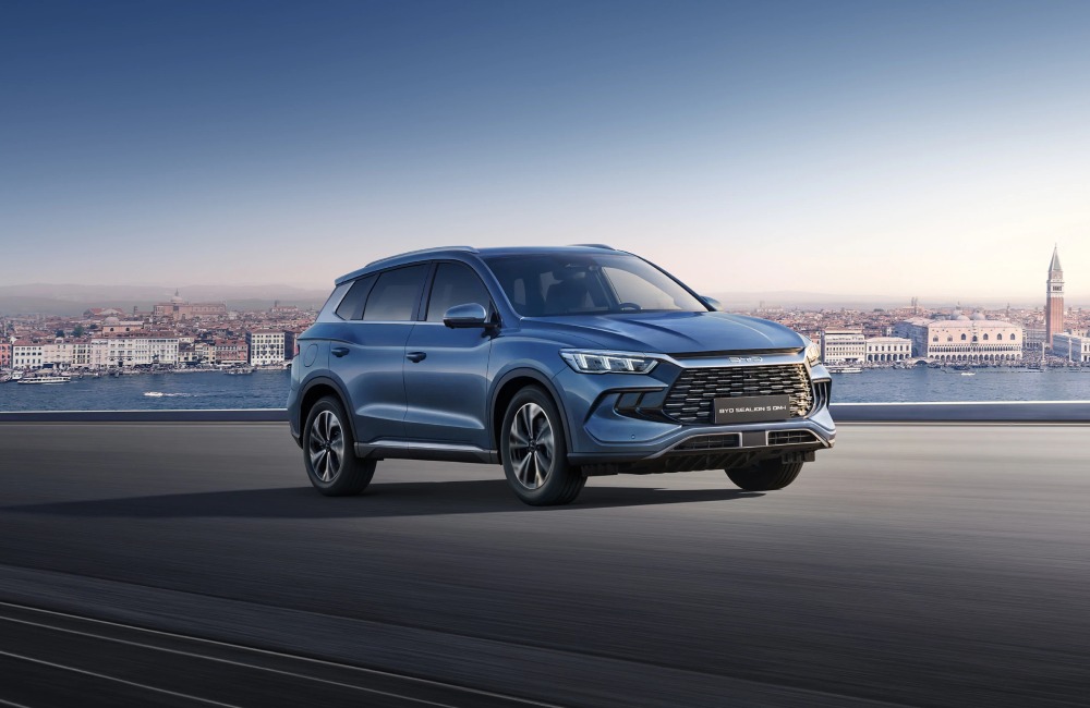 Picturesque image of a BYD SEALION 5 DM-i driving along a tarmac road which overlooks a river with a cityscape behind