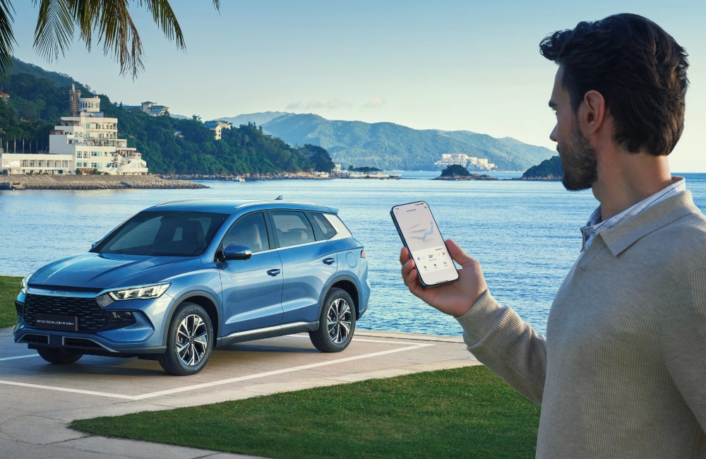 A person utilising an application on his phone to monitor his BYD SEALION 5 DM-i. The car is parked within a square bay, with a lake backdrop behind.