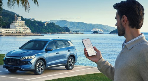 A person utilising an application on his phone to monitor his BYD SEALION 5 DM-i. The car is parked within a square bay, with a lake backdrop behind.