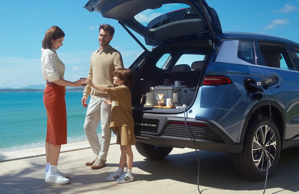 A family consisting of two adults and one child, parked up near the beach and having a drink from a coffee machine powered by the internal battery of their BYD SEALION 5 DM-i.