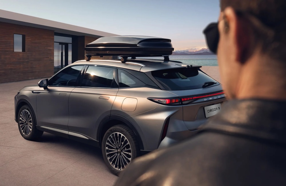 Person with sunglasses looking upon a reared angle of an OMODA 9, the vehicle is parked near a modern wooden house with a scenic ocean and mountain backdrop