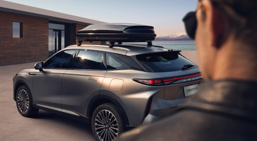 Person with sunglasses looking upon a reared angle of an OMODA 9, the vehicle is parked near a modern wooden house with a scenic ocean and mountain backdrop