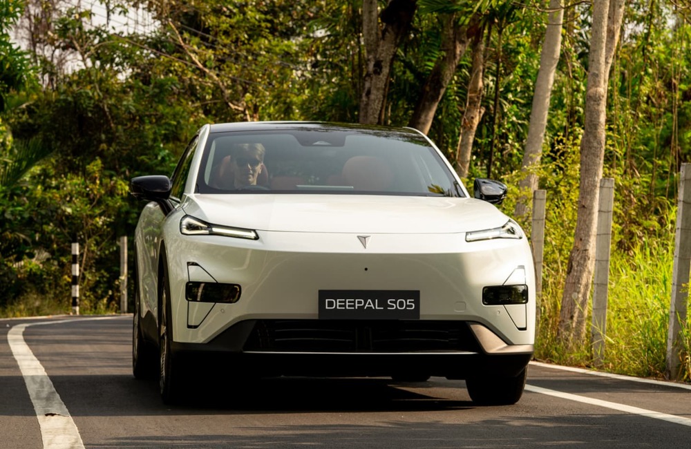 Front view of a Changan Deepal S05 driving down a country road surrounded by trees and brush. The car is white and showing off daytime running lights.