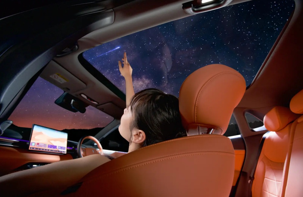Interior shot of a person looking at stars through the panoramic sunroof at night. The interior is orange and lit by the central touchscreen.