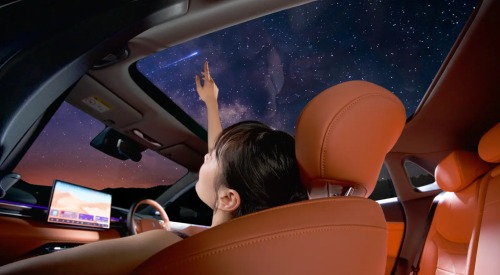 Interior shot of a person looking at stars through the panoramic sunroof at night. The interior is orange and lit by the central touchscreen.